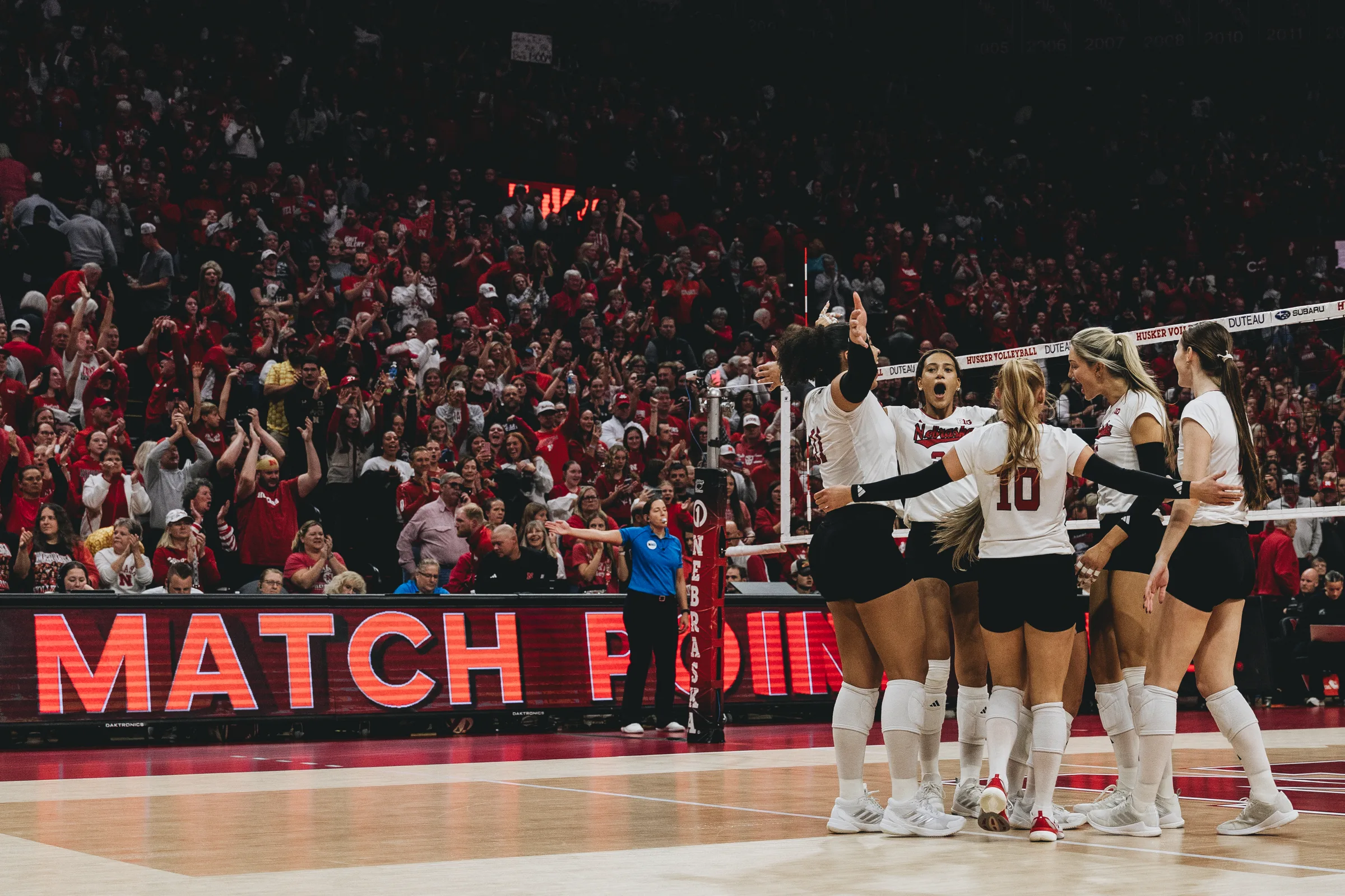 Nebraska Cornhuskers volleyball players celebrate at the net as the MATCH POINT sign blazes on the LED scoreboard and a packed crowd erupts at the Bob Devaney Sports Center in Lincoln, Neb.